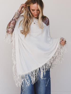 Woman wearing a white fringe poncho over a floral top and jeans on a plain background
