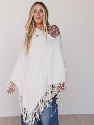 Woman wearing a white poncho with fringe details against a plain background