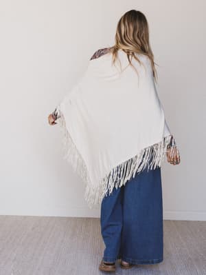 Person wearing a white poncho with fringe details on a plain background