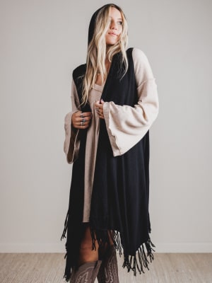 Woman wearing a black and beige hooded vest with fringe details on a plain background