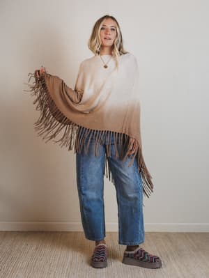 Woman wearing a beige sweater and blue jeans with a brown fringed shawl, standing against a plain wall.