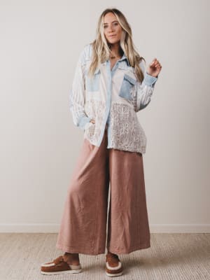 Woman wearing a layered outfit with a lace top, denim jacket, and brown pants against a plain background.