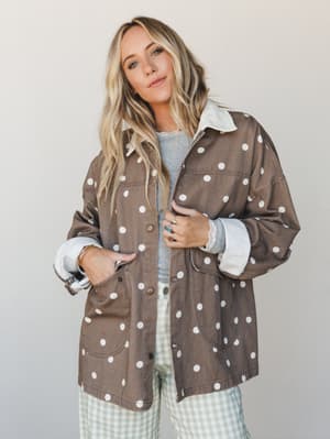 An oversized polka dot chore jacket in mocha brown with a cream contrast collar gets layered over a gray tee and sage gingham pants — the kind of outfit that looks effortless but is definitely a whole mood.