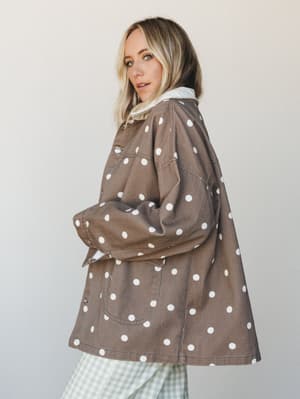 A blonde model shot in three-quarter profile against a soft neutral studio wall, wearing an oversized polka dot chore jacket in warm mocha paired with sage green gingham wide-leg pants — the combo feels effortlessly collected, like she put it togethe