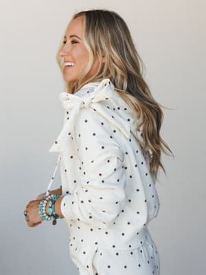 A smiling model shot in profile shows off the back bow detail of a cream polka dot hoodie — the kind of piece that feels playful and intentional all at once, styled with stacked turquoise bracelets for that easy, personal-touch energy.