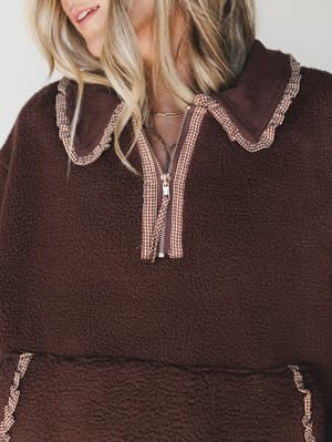 A close-up of a rich chocolate sherpa pullover with gingham-trimmed ruffle detailing along the collar and zipper placket — the kind of cozy piece that makes you want to immediately cancel plans and stay home.