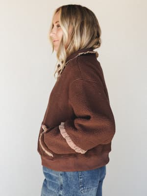 A side-profile shot of a model in a rich chocolate brown sherpa pullover with gingham ruffle trim at the collar and sleeves, paired with medium-wash jeans — cozy and quietly interesting.