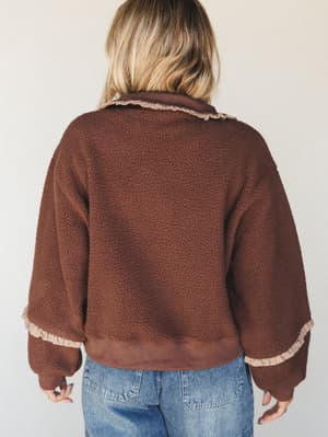 Shot from behind, this cozy sherpa pullover in deep chocolate brown has a sweet surprise — delicate gingham ruffle trim at the collar and cuffs that keeps it from feeling too casual, paired back with relaxed light-wash jeans.