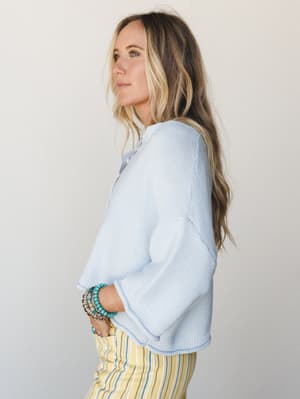 A side-profile shot of a model in a soft powder blue cropped knit sweater layered over a collared shirt, paired with yellow striped pants and a stack of turquoise and mixed bead bracelets — easy, warm-weather layering with a relaxed boho feel.