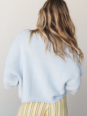 A back-facing shot of a cropped baby blue knit sweater with relaxed short sleeves, paired with yellow striped bottoms — soft, breezy, and effortlessly warm-weather ready.