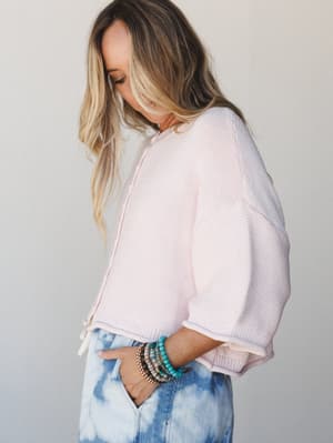 A soft, dreamy side profile of a model in a blush pink cropped knit sweater and tie-dye jeans, her wrist stacked with beaded bracelets and her hand tucked into her pocket — the kind of outfit that feels effortless but totally intentional.
