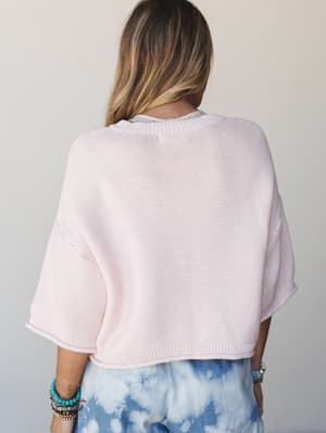 A back-view shot of a boxy cropped knit sweater in the softest blush pink, layered over a printed top and paired with tie-dye bottoms — the kind of effortless combo that looks accidental but really isn't.
