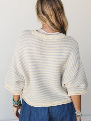 A back-facing shot of a boxy striped knit sweater in cream and soft periwinkle, layered over a denim skirt and styled with stacked turquoise and wood bead bracelets — the kind of effortless outfit that looks like it happened without trying.