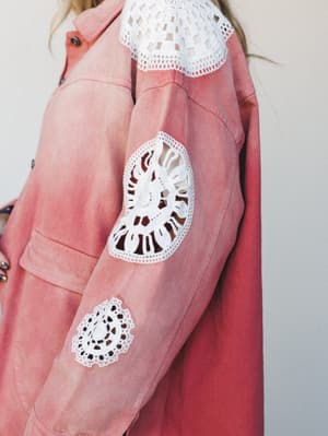 A close-up detail shot of a washed rust denim jacket layered over a white crochet top, with standout macramé appliqués scattered across the sleeve — the kind of piece that makes you stop mid-scroll.
