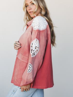 A washed rust denim jacket with standout white crochet medallion patches at the shoulder, elbow, and cuff — worn over light jeans with stacked rings, shot from the side in soft natural light.