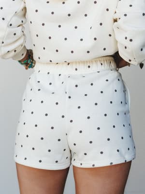 A back-view close-up of a cream polka dot matching set — cropped sweatshirt and pull-on shorts — styled with layered boho bracelets, giving off that effortlessly cozy, staying-in-but-make-it-cute energy.