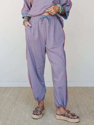 A relaxed striped jogger pant in dusty blue with red pinstripes, styled as a matching set with a coordinating sweatshirt, platform espadrille sandals, and a stack of turquoise and beaded bracelets — the kind of outfit that looks effortless but clearl