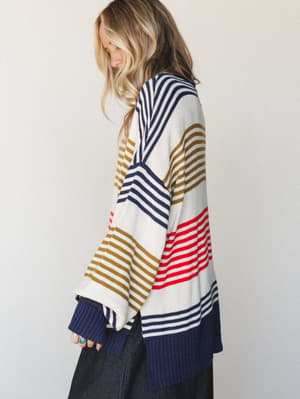 A side-profile shot of a model in an oversized multi-stripe knit sweater with bold color-blocked stripe panels in navy, red, and olive over cream, paired with wide-leg dark denim — the kind of sweater that looks like it has a whole personality.