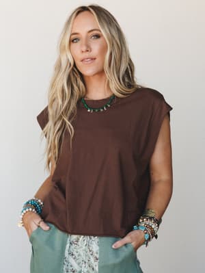 A relaxed, sleeveless muscle-tee in deep chocolate brown styled with sage wide-leg pants, stacked boho bracelets, and a turquoise letter bead necklace — effortless and personal, like an outfit that just happened to look that good.