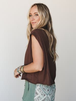 A model glances back over her shoulder in a cropped dark chocolate tee with sleeveless styling, paired with a sage and floral patchwork bottom — the stacked boho bracelets and easy, lived-in vibe doing all the right things.