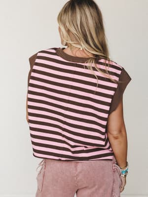 Shot from behind, a blonde model wears a boxy striped tee in pink and chocolate brown with solid brown cap sleeves, paired with dusty rose acid-wash cargo pants and a stack of beaded bracelets — casual and a little retro, like something you'd grab of