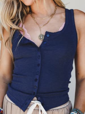 A close-up mid-shot of a navy button-front tank top layered over a blush lace bralette, styled with a taupe drawstring skirt and layered gold necklaces — casual and personal, the kind of outfit that looks like it came together without trying.