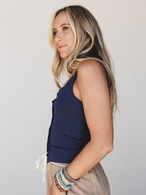 A model shot in three-quarter profile wearing a fitted navy button-front tank with a side tie detail, layered with stacked beaded bracelets and paired with soft taupe bottoms — easy, personal, and quietly put-together.
