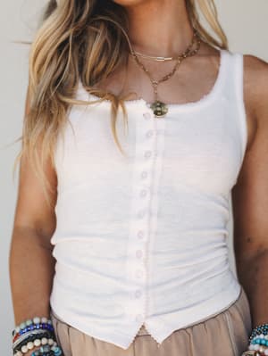 A ribbed button-front tank top in soft white-blush is styled with a flowy taupe skirt, stacked beaded bracelets, and layered gold necklaces — the kind of effortless warm-weather look that feels as good as it looks.