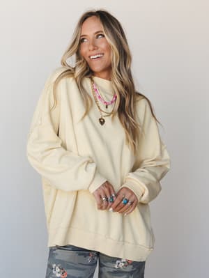 An oversized cream sweatshirt styled with layered gold and pink beaded necklaces, stacked silver and turquoise rings, and floral camo pants peeking through at the bottom — the kind of effortless, personality-forward outfit that looks like it took zer