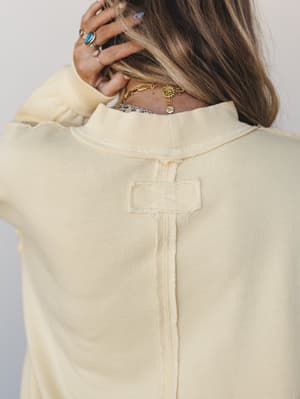 A back-detail shot of a soft cream sweatshirt with exposed seaming and a cross-stitch patch detail at the center back, styled with layered gold and silver necklaces and stacked turquoise rings — the kind of piece that looks intentional without trying
