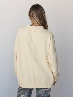 A back-view shot of an oversized cream sweatshirt with exposed seam detailing down the center back, paired with floral camo pants — relaxed and lived-in with just enough personality.