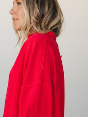 A close-up side profile shot of a bold red top with layered flutter sleeves, the raw-edge seam detail doing quiet, interesting work against a clean studio backdrop.