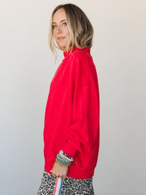 A model shot in profile wearing an oversized red quarter-zip pullover layered over a leopard print bottom, styled with a stack of colorful beaded bracelets — casual and expressive with just enough personality to make a plain sweatshirt feel intention