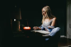 A woman curled up in a dim, candlelit room with a laptop and open journal, wearing a grey lace-trim cami layered under a chunky knit cardigan — the whole scene feels like a quiet Sunday evening with nowhere to be.