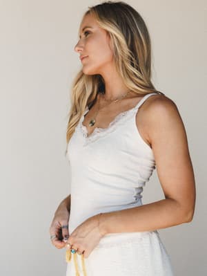 A soft, sun-warmed shot of a white lace-trim cami styled with layered gold jewelry and a peek of a textured white skirt — effortless and feminine, like she just decided to look this good.