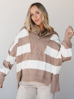 An oversized quarter-zip pullover in warm mocha and cream wide stripes, styled with cream corduroy pants against a soft neutral studio backdrop — the kind of effortless cozy outfit you'd actually wear on a Saturday and still feel put-together.