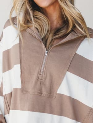 A close-up shot of a quarter-zip pullover in wide mocha and cream stripes, with a ribbed placket and silver zipper — the kind of top that looks equally good on a coffee run or a cozy weekend wherever.
