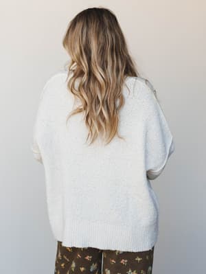 Shot from behind, a model wears an oversized ivory knit sweater with a relaxed, boxy silhouette paired with dark floral wide-leg pants — the combination has that effortless, cozy-meets-expressive energy.