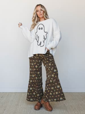 A relaxed, full-body studio shot of a model in an oversized ivory knit sweater with a checkered ghost graphic paired with dark brown floral wide-leg cargo pants and studded clogs — the kind of outfit that makes Halloween feel cozy and personal rather
