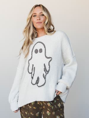 An oversized cream knit sweater with a charming gingham-outlined ghost graphic is styled with dark floral wide-leg pants — the kind of cozy, personality-forward outfit that makes October feel like a whole personality.