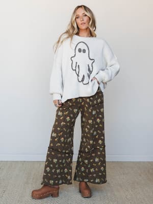 A cream knit sweater with a playful checkered ghost graphic is paired with wide-leg dark floral pants and brown studded clogs — the kind of outfit that makes Halloween feel like a personality, not a costume.