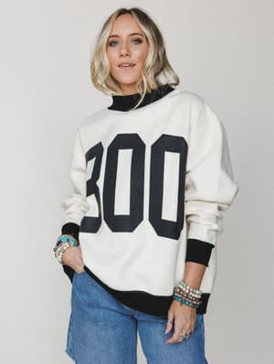 A cream oversized graphic sweatshirt with bold 'BOO' lettering and black contrast trim, styled with wide-leg jeans and stacked beaded bracelets — casual Halloween dressing that doesn't take itself too seriously.