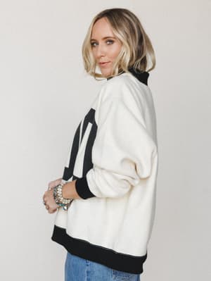 A relaxed, side-profile shot of a model in an oversized cream sweatshirt with bold black graphic lettering, styled with stacked beaded bracelets and the hint of blue denim — the kind of effortless outfit you'd wear to a farmers market and get complim