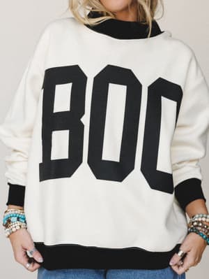 An oversized cream sweatshirt with bold black 'BOO' varsity lettering and contrast ribbed trim, styled with stacked boho bracelets and a peek of denim — casual, playful, and very much the sweater you grab on the way out the door in October.