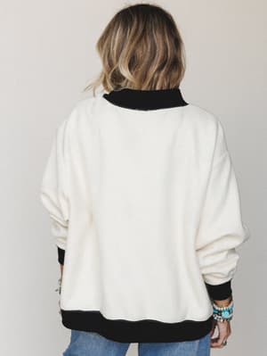 A back-view shot of a cream fleece pullover with black contrast ribbed trim at the collar, cuffs, and hem — styled with denim and a stack of turquoise and gold bracelets for that effortlessly collected everyday look.