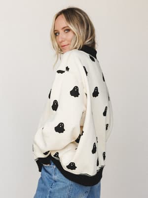 A blonde model looks over her shoulder wearing an oversized cream sweatshirt covered in black ghost print, paired with light wash denim — casual and a little witchy in the best way.