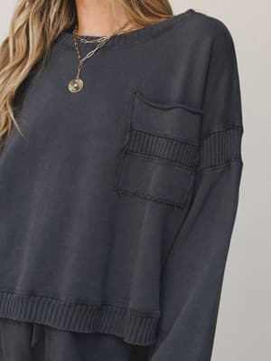A close-up of a washed charcoal pullover sweatshirt with raw-edge chest pocket and ribbed sleeve detailing, styled with a layered gold chain necklace — the kind of top that does the heavy lifting on a do-nothing day.
