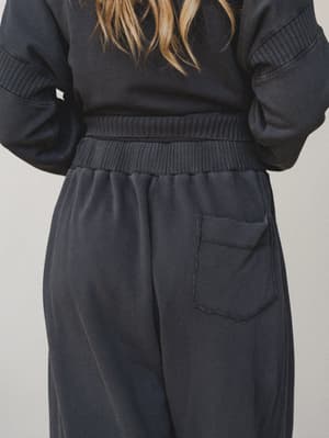 A back-view detail shot of a matching charcoal sweat set — the relaxed joggers and ribbed-paneled sweatshirt feel intentionally worn-in, with raw-edge pocket stitching and a wide elastic waistband that give the set a laid-back, lived-in quality.