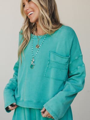 A relaxed cropped sweatshirt in washed turquoise green styled with layered gold and blue necklaces — the kind of effortless top that looks just as good on a coffee run as it does paired with matching bottoms on a slow Sunday.
