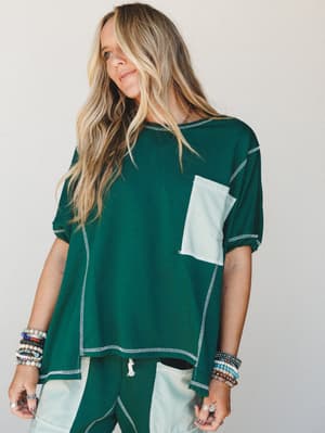 A relaxed, mix-fabric top in deep forest green with contrast stripe pocket and exposed stitching, styled with matching wide-leg pants and stacked boho bracelets — the kind of set that blurs the line between staying in and going out.